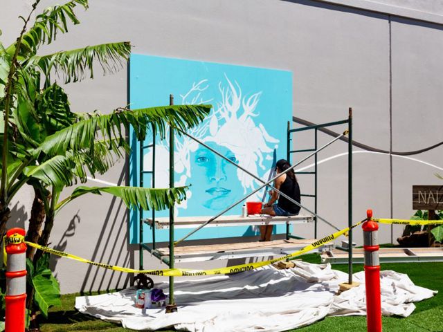 An artist sits on scaffolding painting a mural of a woman with marine organisms for hair and a bright turquoise background.