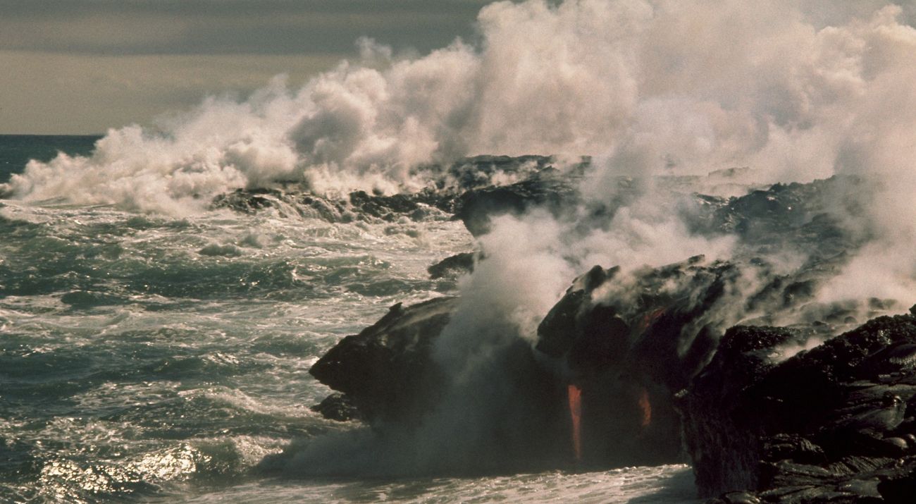 Strong waves crash up against hot volcanic rock and molten lava, causing steam.