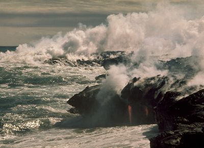 Lava pours off a rocky cliff edge into wavy ocean waters, and huge clouds of steam blow in the air above.
