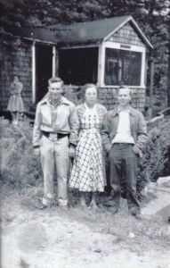 Black-and-white image of two young man standing with an older woman standing by a cabin, dressed in the style of the 1940s.