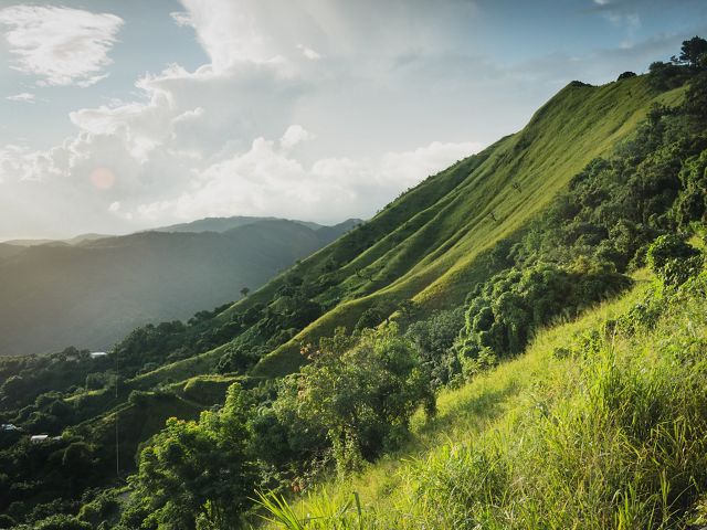 The verdant green slope of a steep mountain set against a blue sky with puffy white clouds.