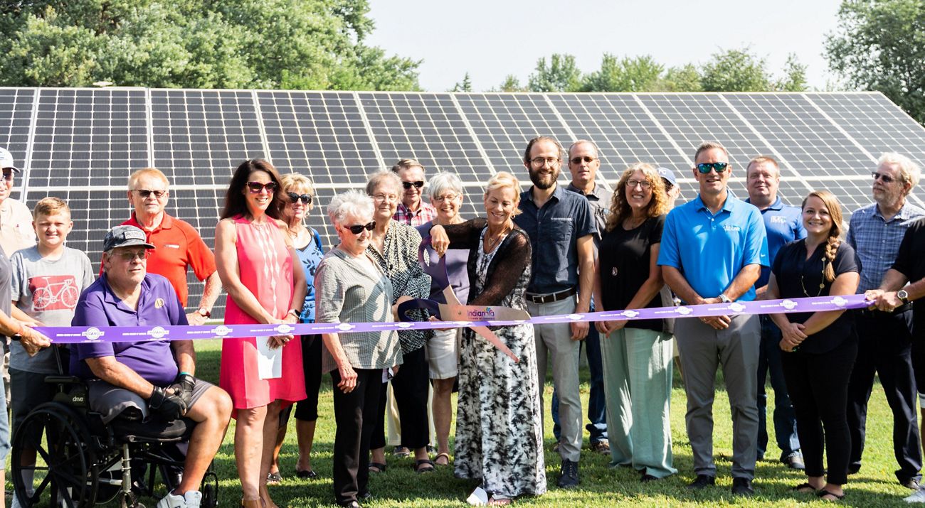A group of more than a dozen adults gather in front of solar panels, while a person in the front center uses a giant pair of scissors to cut a purple ribbon that extends across the group.