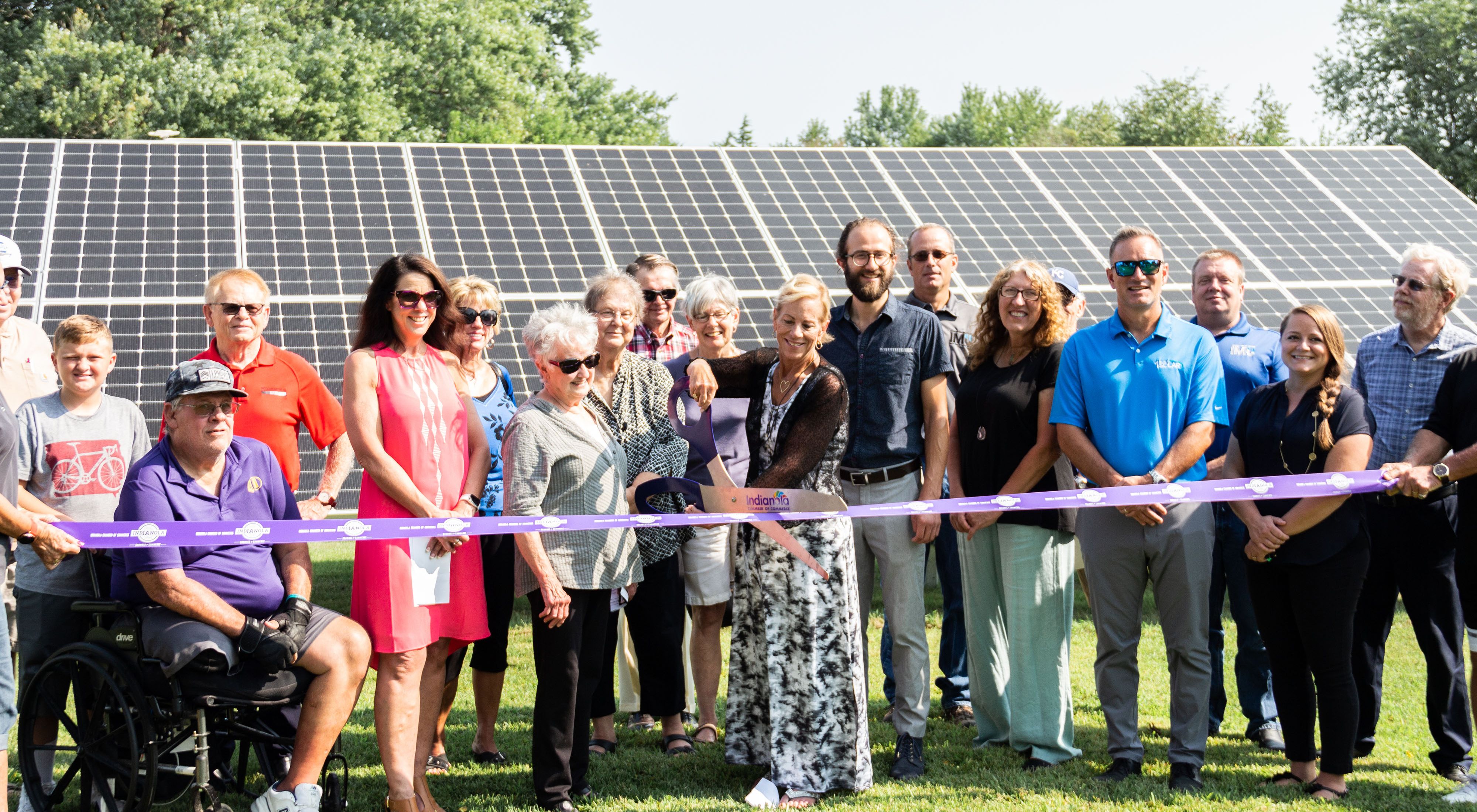 A group of more than a dozen adults gather in front of solar panels, while a person in the front center uses a giant pair of scissors to cut a purple ribbon that extends across the group.
