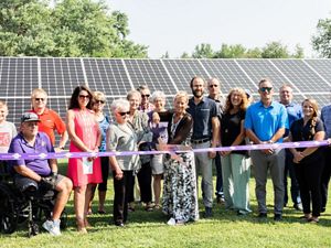 A group of more than a dozen adults stand in front of solar panels, while a person in the front center uses a giant pair of scissors to cut a purple ribbon that extends across the group.