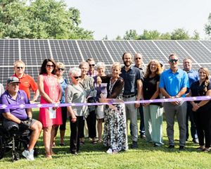 A group of more than a dozen adults stand in front of solar panels, while a person in the front center uses a giant pair of scissors to cut a purple ribbon that extends across the group.