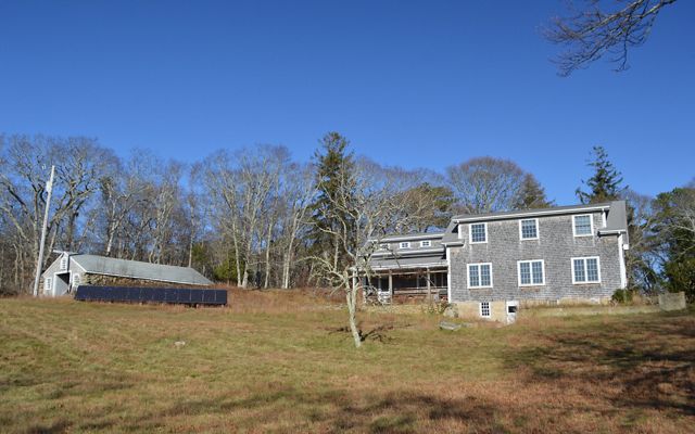 A gray shingled house sits on a grassy hill with a solar panel array to the left of it and trees and blue sky in the background.