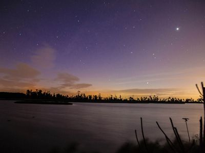 The sky turns purple and stars appear over an orange horizon as the sun sets over a wooded island and open body of water.