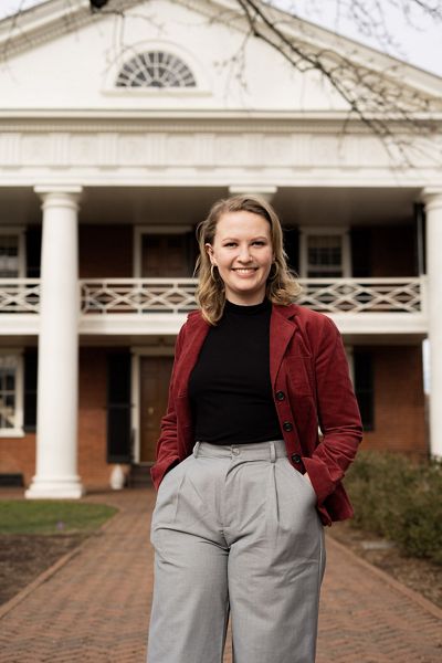A woman wearing a red jacket smiles in front of an historic home.