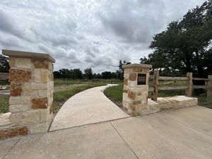 Stone entry pillars and a sign reading Honey Creek State Natural Area frame a newly opened trailhead and paved path leading into grassland and oak trees beneath a cloudy sky.