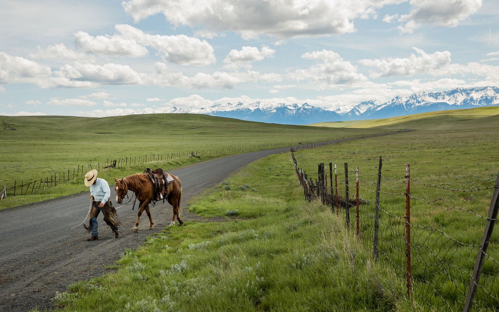 A man in a cowboy hat walks in front of a horse along a dirt road that cuts through a large, green, grassy prairie, with a mountain range in the background.