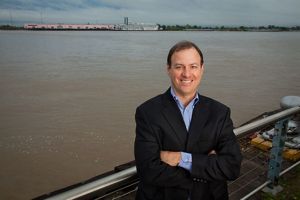 A smiling man in a blazer stands in front of a river.