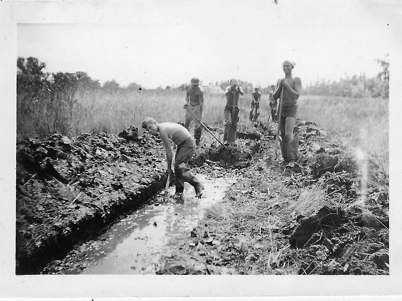 Black and white photo from 1935. Five men holding shovels stand at the end of a wide ditch filled ankle deep with water. One man is in the ditch bent over digging.
