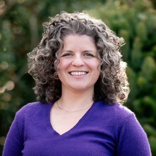 Headshot of Emma Gildesgame in front of trees. She wears a purple sweater and has curly black and grey hair.