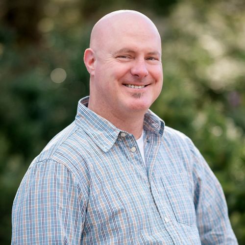 Headshot of Rene Wendell in a blue plaid shirt with greenery in the background.