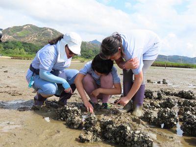 A teacher and two students squat to look at specimens in the mud.