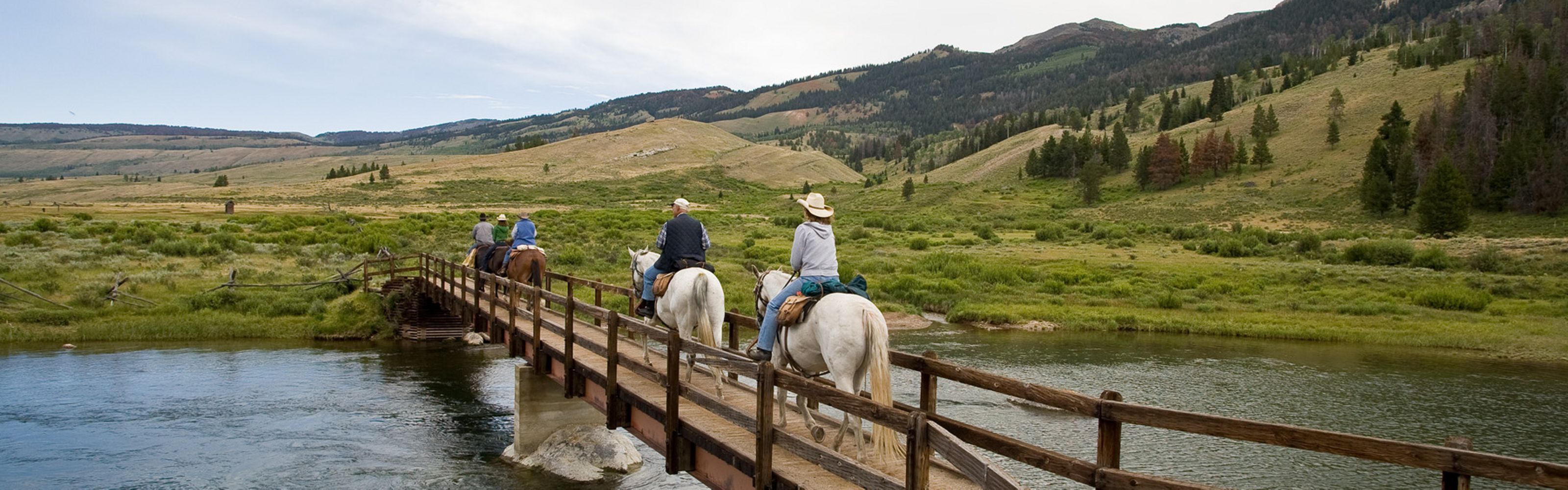 Five people on horseback cross a narrow wooden bridge over a stream, with green rolling hills and mountains in the background.