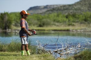 A young boy stands on the edge of a blue pond fishing with a rod.