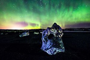 A crystal-clear block of ice is reflected in the foreground, and a shockingly vibrant aurora borealis fills the sky in the background.
