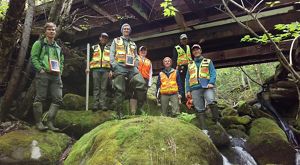 People in safety vests pose on rocks in a stream bed with a steel bridge overhead.