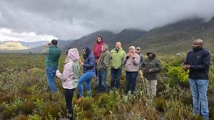 A group of 11 adults stands together wearing different colored jackets in the middle of a mountainous and foggy area. 