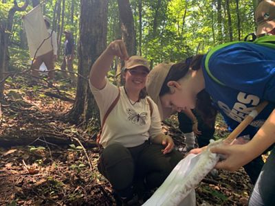A group of three students conduct fieldwork in a forest. A smiling student holds up a small plastic sample bottle containing a tick.
