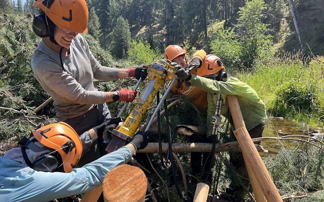 A group of four wearing orange hard hats hold up a yellow piece of equiptment.