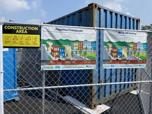 Two signs with graphics and text hang on a metal link fence around a parking lot.