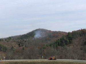 Smoke from a controlled burn rises above a green forested hill as cars drive by on the interstate highway that runs along the edge of the forest.
