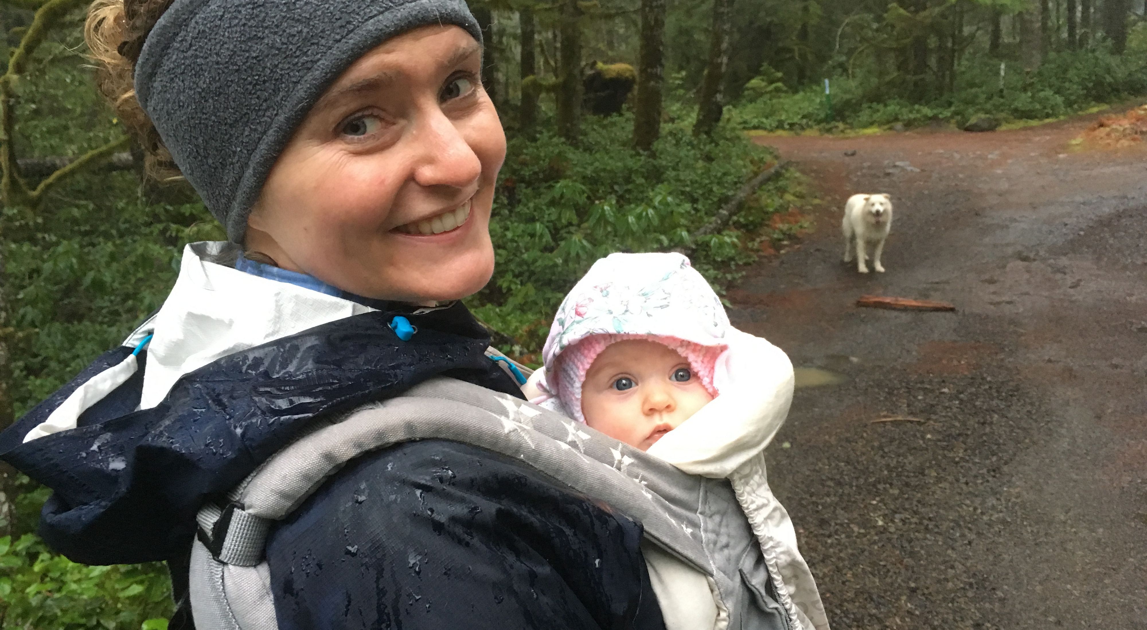 A woman holds a baby and smiles at the camera as they walk through the woods; a white dog stands on a dirt trail in the background.