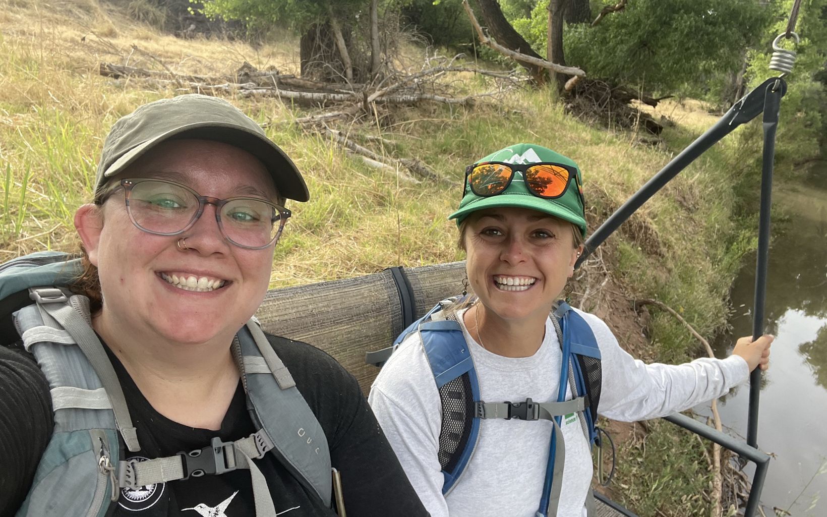 Two TNC staff members, Taylor Hansen and Jessie Pearl, sit next to a river and smile at the camera.