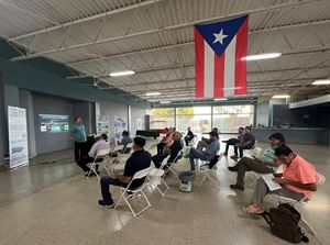 people sit under a Puerto Rican flag in a lecture hall setting listening to a speaker.
