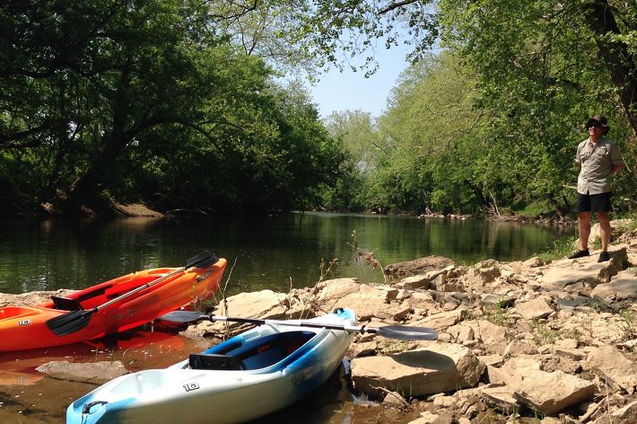 A man stands on the rock lined shore of a narrow river. Two kayaks, orange and white, are pulled up onto the rocks in the foreground. The river is thickly lined with trees that overhang the water.