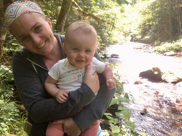 Julia Amato holds her baby and they both smile at the camera as they stand next to a rushing stream in a forest.