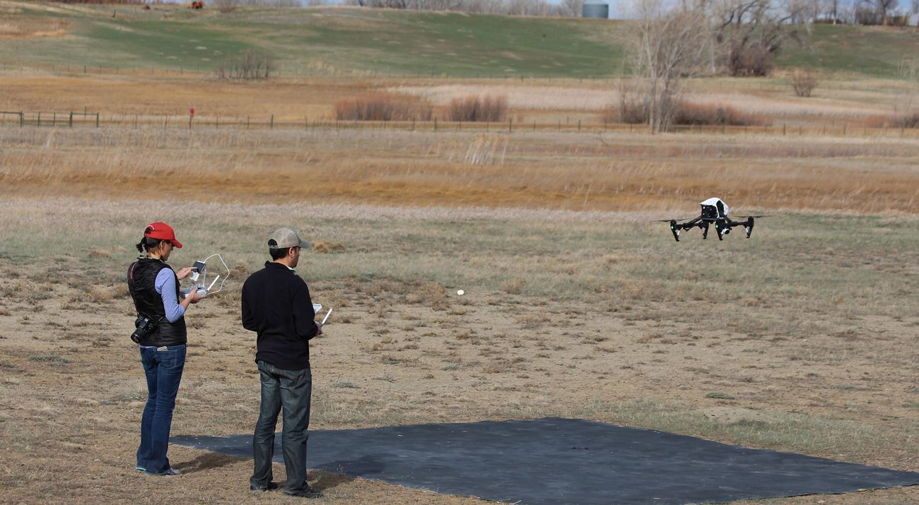 Two people stand in a large grassy field and fly a drone.