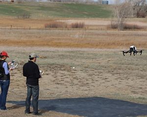 Two people stand in a large grassy field and fly a drone.