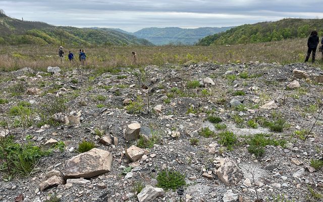A field of compacted dirt and rubble in a former mineland gives way to a grassy restored area ringed by tree covered hills.