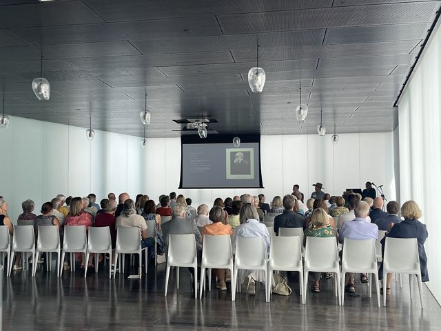 Rows of people sit in white chairs facing a presenter who is delivering a lecture.