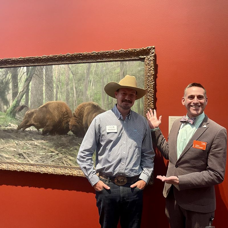 Two men, one wearing a tan cowboy hat and the other a bowtie, smile in front of a painting of two bison locking horns.