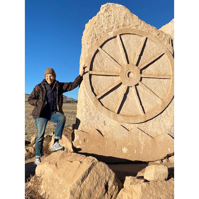 A woman leans against a large stone with a wheel shape on it.