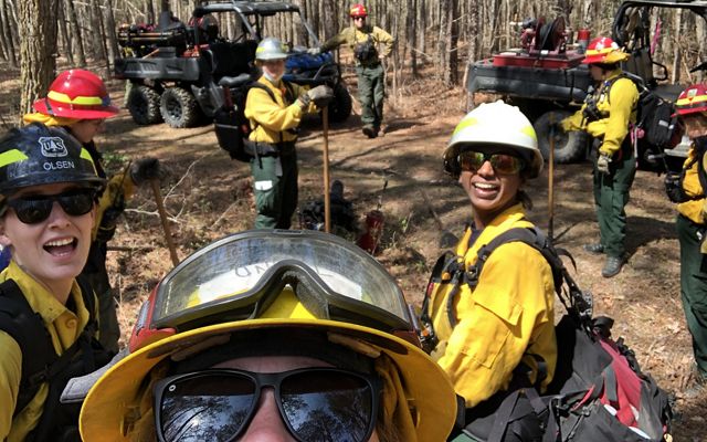 A woman holds up a camera to capture a group selfie of the six women standing behind her during a fire learning exchange. They are standing in a forest next to two ATVs.