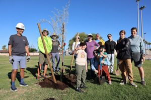 A group of adults and children stand around a newly planted tree in a sunny park.