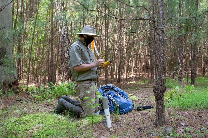 A man sits on his knees recording data into a hand held tablet that it attached to a length of tube in the ground.