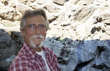 Portrait of a older man with graying hair and white beard. He is standing in front of a tall rock face.