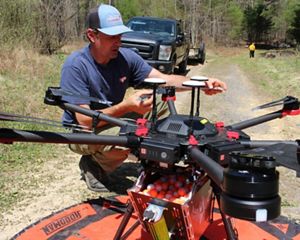 A man crouches on the ground next to a large drone, readying it for deployment on a controlled burn. The drone is sitting on a round orange landing pad.