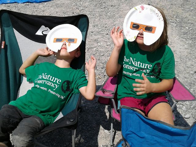 Two children wear homemade solar eclipse glasses made from paper plates.