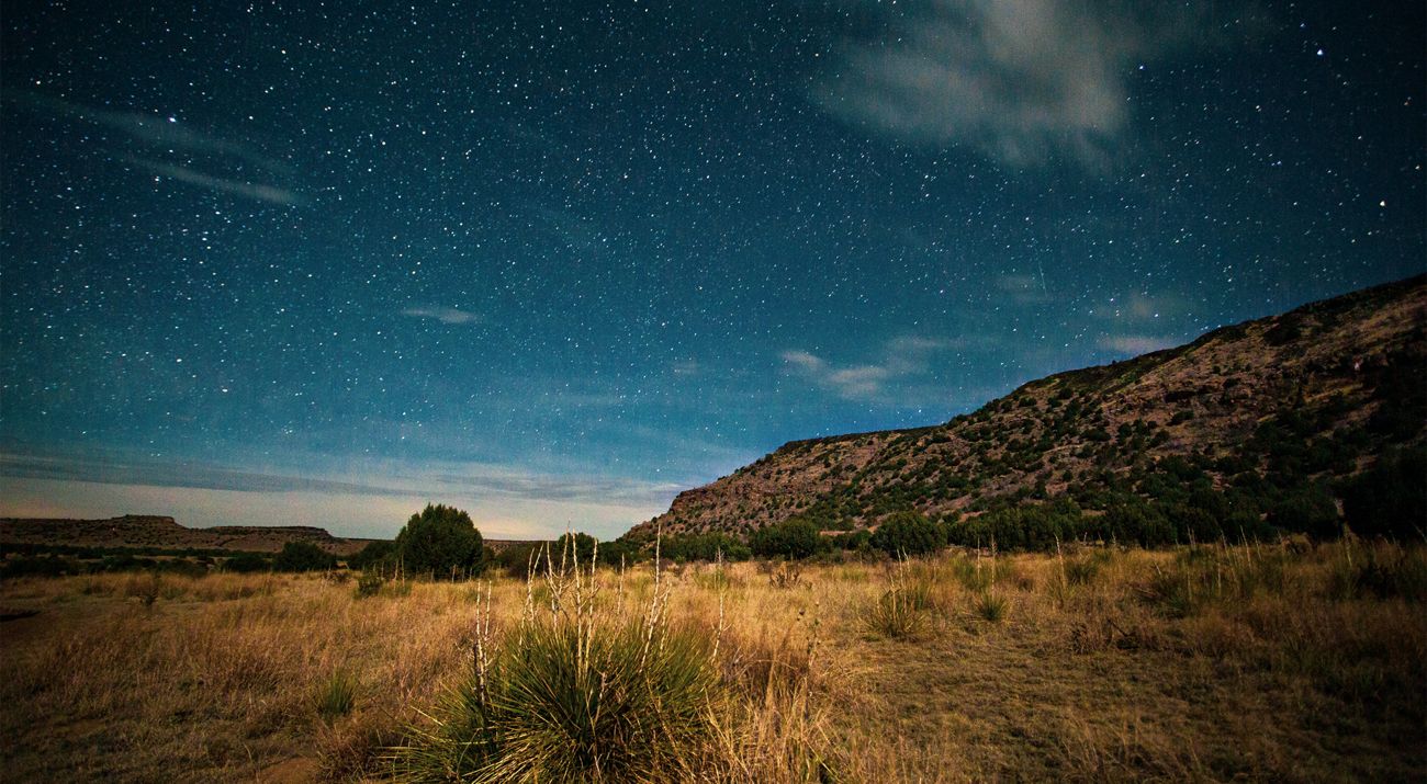 A scrubby landscape and hills lay under the emerging stars as the sun sets at Black Mesa State Park and Nature Preserve.