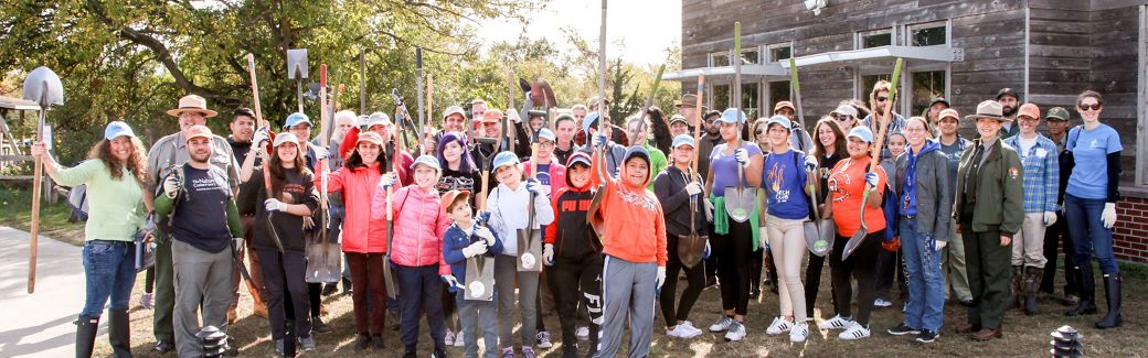 A large group of people standing outside of a building on the right hand side of the photo with green trees featured in background. Most of the people are holding gardening tools and smiling.