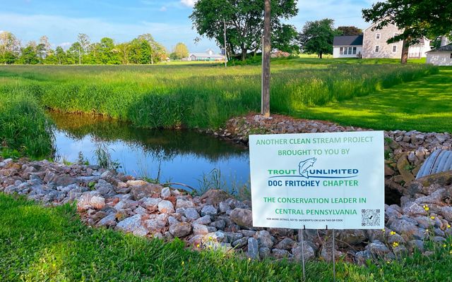 A sign about healthy streams sits in front a small body of water surrounded by green grass.