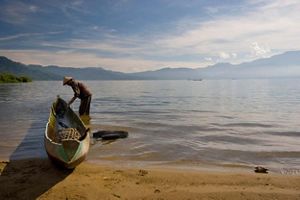 fisherman in conical hat cleans wooden boat at a lakeshore.