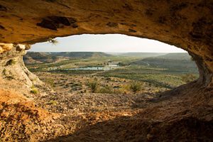 A balanced rock with a circular hole in the center reveals desert mesas and a small pond.